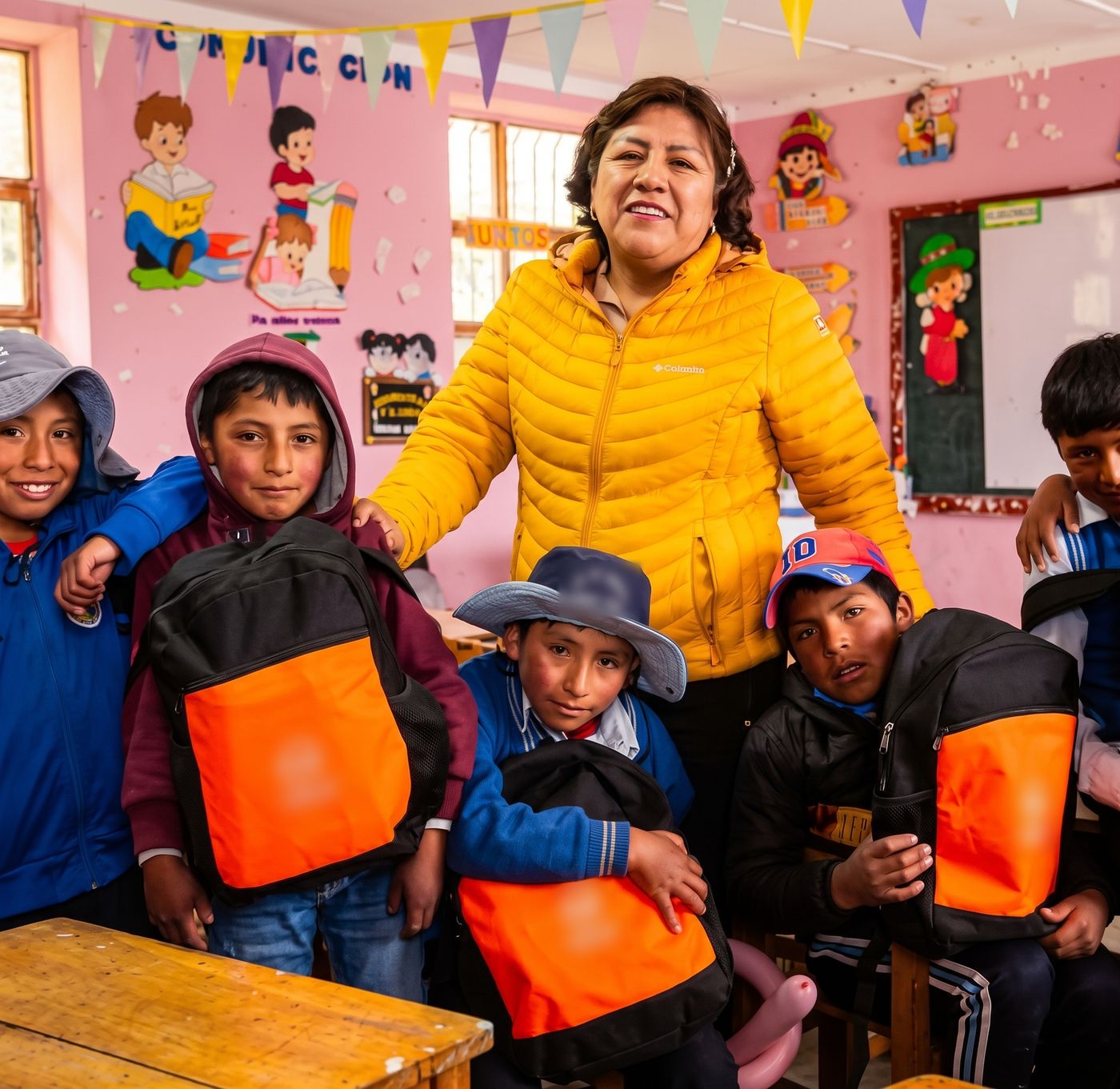 Una docente y cinco niños en un aula; los niños sostienen mochilas negras y naranjas de un kit escolar.